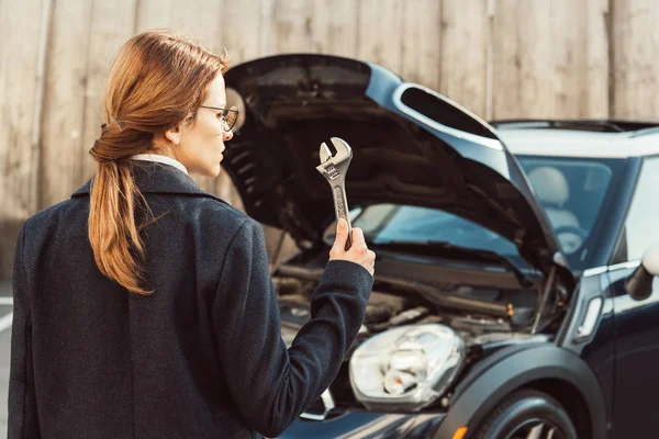 Selective focus of woman in coat standing with wrench near car at city street — Stock Photo