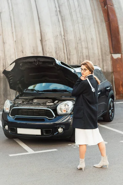 Frau in Mantel und Brille posiert mit Schraubenschlüssel neben Auto mit geöffneter Motorhaube auf Stadtstraße — Stockfoto
