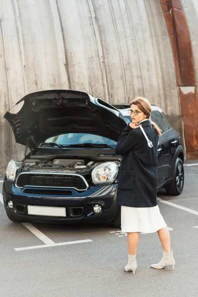 Female woman in coat and eyeglasses posing with spanner near car with opened hood at urban street — Stock Photo