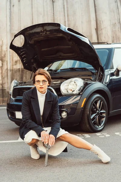 Femme adulte à la mode en manteau et lunettes posant avec clé près de la voiture avec capuche ouverte dans la rue urbaine — Photo de stock