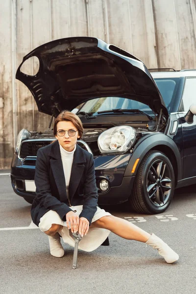 Mujer adulta de moda en abrigo y anteojos posando con llave cerca del coche con capucha abierta en la calle urbana - foto de stock