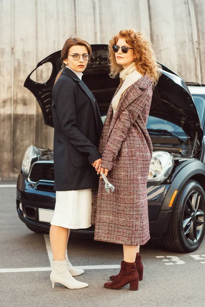 Dos elegantes modelos femeninos en abrigos que sostienen la llave inglesa cerca del coche con capucha abierta en la calle de la ciudad - foto de stock