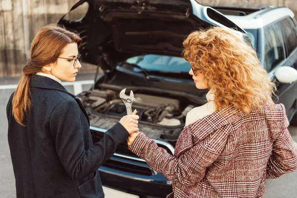 High angle view of curly ginger woman giving wrench to female friend in coat near car with opened hood at city street — Stock Photo