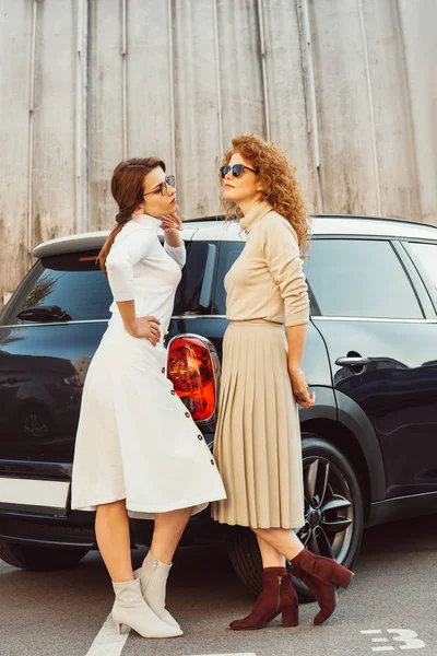 Modelos femeninos adultos con estilo en gafas de sol y faldas posando cerca de coche en la calle urbana - foto de stock