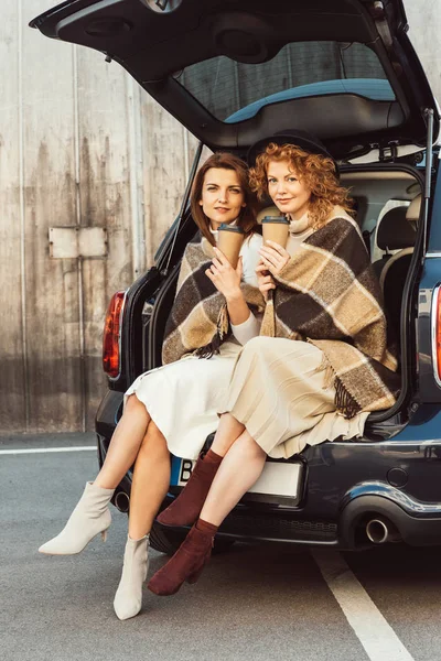 Atractivas amigas envueltas en mantas sosteniendo tazas de café desechables y sentadas en el maletero del coche en la calle de la ciudad - foto de stock