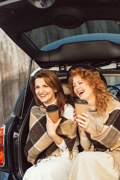 Selective focus of laughing women wrapped in blankets holding disposable coffee cups and sitting in car trunk at street — Stock Photo