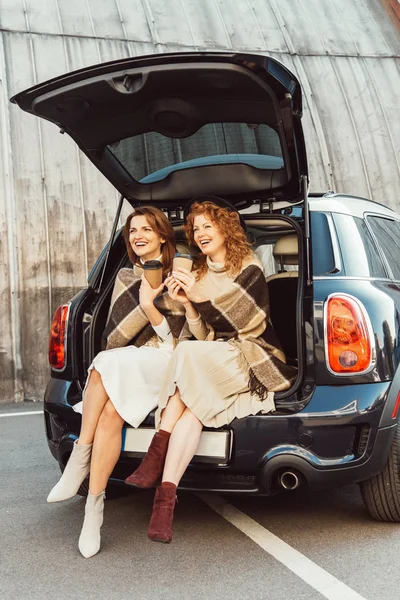 Laughing adult women wrapped in blankets holding disposable coffee cups and sitting in car trunk at city street — Stock Photo