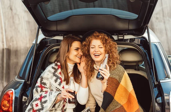 Side view of smiling woman with soda bottle whispering to laughing redhead female friend in car trunk at city street — Stock Photo
