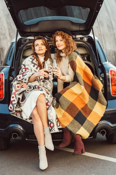 Mujeres elegantes atractivas envueltas en mantas que sostienen botellas de refresco sentadas en el maletero del coche en la calle urbana - foto de stock