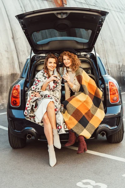 Веселі друзі-жінки, загорнуті в ковдри, тримають пляшки содової, сидячи в багажнику автомобіля на міській вулиці — стокове фото