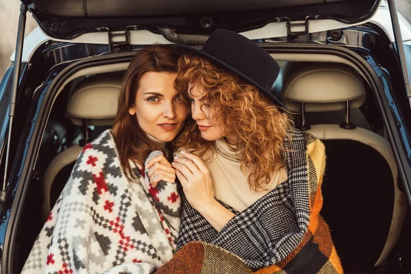 Portrait of pretty female friends wrapped in blankets sitting in car trunk at city street — Stock Photo