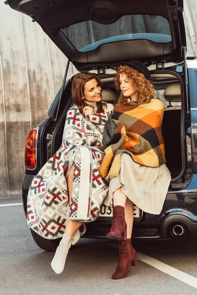 Happy adult women wrapped in blankets sitting in car trunk at urban street — Stock Photo