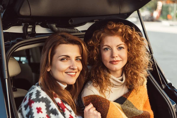 Portrait of attractive women wrapped in blankets sitting in car trunk at street — Stock Photo