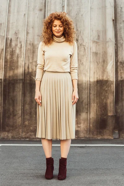 Cheerful curly redhead woman in stylish beige turtle neck and skirt posing at city street — Stock Photo