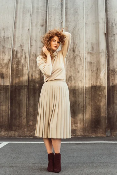 Stylish curly redhead woman in beige turtle neck and skirt posing at city street — Stock Photo