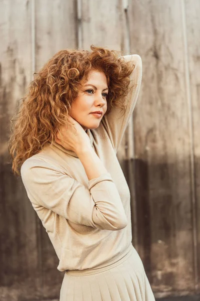 Portrait of attractive curly redhead woman in beige turtle neck posing at city street — Stock Photo