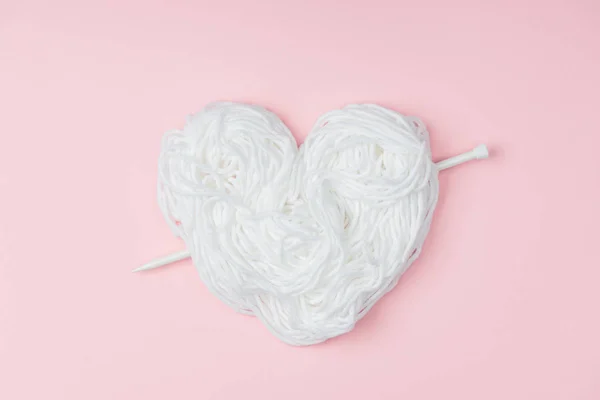 Top view of yarn arranged in heart symbol and knitting needle on pink backdrop — Stock Photo