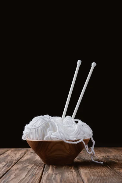 Vista ravvicinata di filati bianchi in ciotola e ferri da maglia su una superficie di legno su sfondo nero — Foto stock