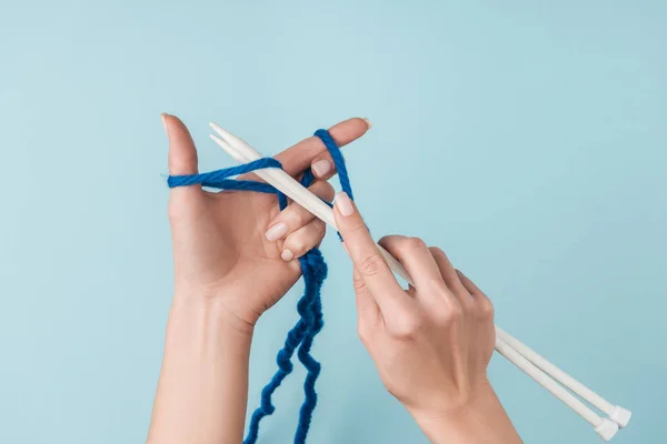 Partial view of woman with blue yarn and white knitting needles knitting on blue backdrop — Stock Photo