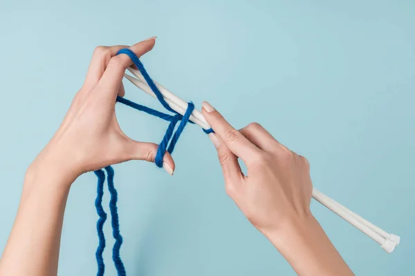 Partial view of woman with blue yarn and white knitting needles knitting on blue backdrop — Stock Photo