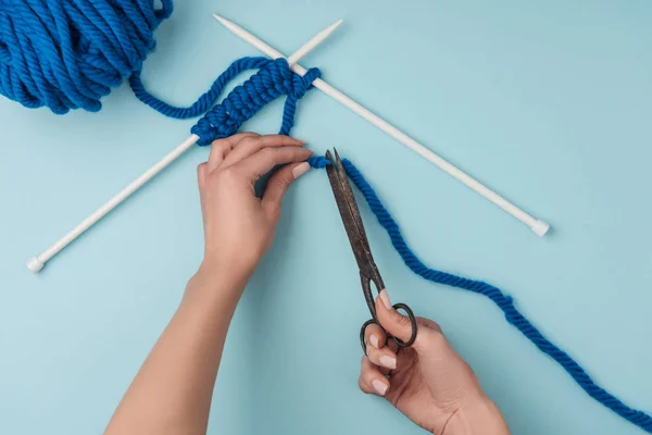 Visão parcial da mulher fio de corte com scissros no fundo azul com fios e agulhas de tricô — Fotografia de Stock
