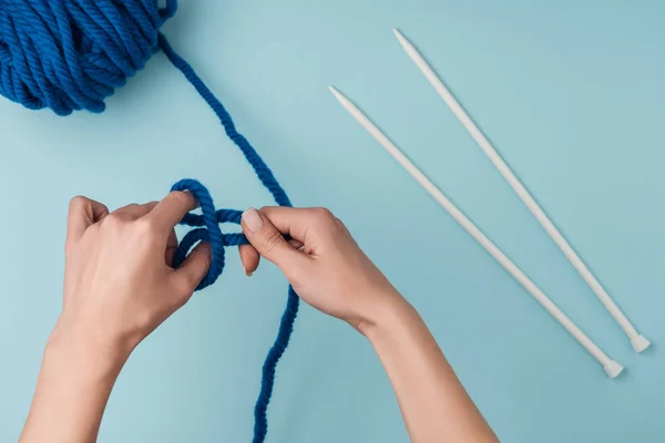 Partial view of woman with blue yarn and white knitting needles knitting on blue backdrop — Stock Photo