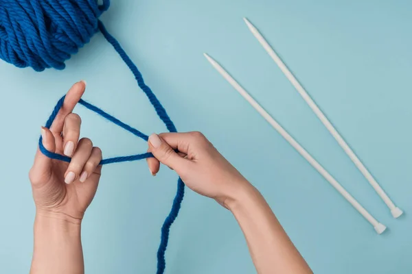 Partial view of woman with blue yarn and white knitting needles knitting on blue backdrop — Stock Photo