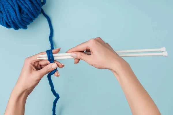 Partial view of woman with blue yarn and white knitting needles knitting on blue backdrop — Stock Photo