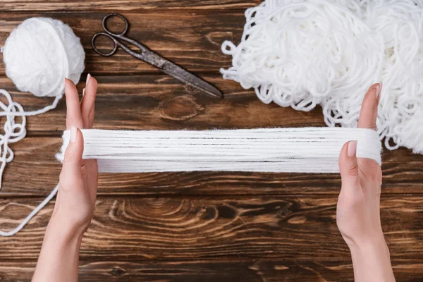 Partial view of woman holding white yarn for knitting on hands on wooden tabletop with scissors — Stock Photo