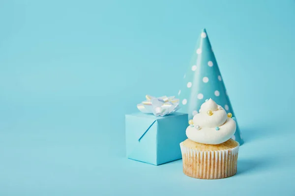 Sombrero de fiesta, caja de regalo y delicioso cupcake sobre fondo azul - foto de stock