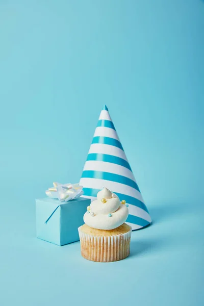Chapéu de festa, caixa de presente e cupcake saboroso com polvilhas de açúcar no fundo azul — Fotografia de Stock