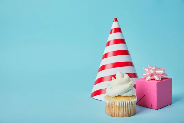 Sombrero de fiesta rojo blanco, caja de regalo rosa y delicioso cupcake sobre fondo azul - foto de stock