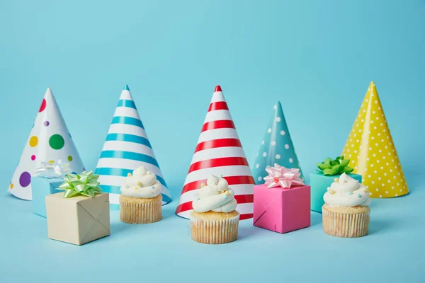 Chapéus coloridos, presentes e deliciosos cupcakes no fundo azul — Fotografia de Stock