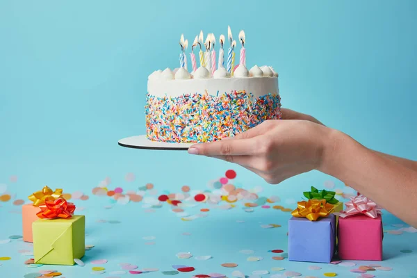 Vista recortada de la mujer sosteniendo pastel de cumpleaños con velas sobre fondo azul con confeti y regalos - foto de stock