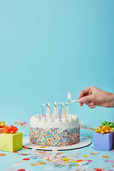 Partial view of woman lighting birthday cake on blue background with gifts and confetti — Stock Photo