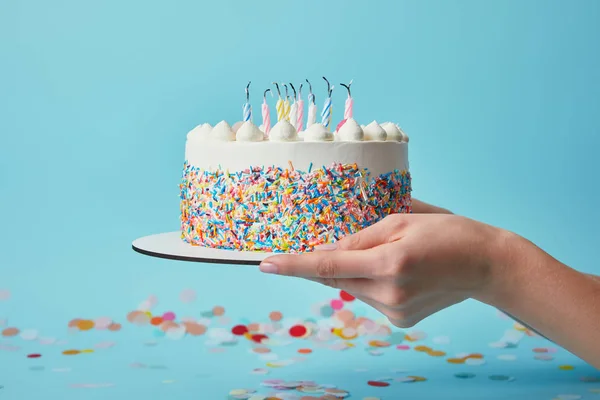 Vista parcial de la mujer sosteniendo pastel de cumpleaños con velas sobre fondo azul con confeti - foto de stock