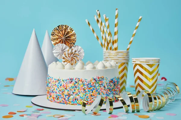 Cake, party hats, plactic cups, golden paper streamer  and drinking straws on blue background with confetti — Stock Photo