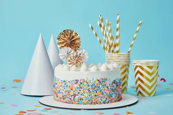 Delicious cake, plactic cups, party hats and drinking straws on blue background with confetti — Stock Photo