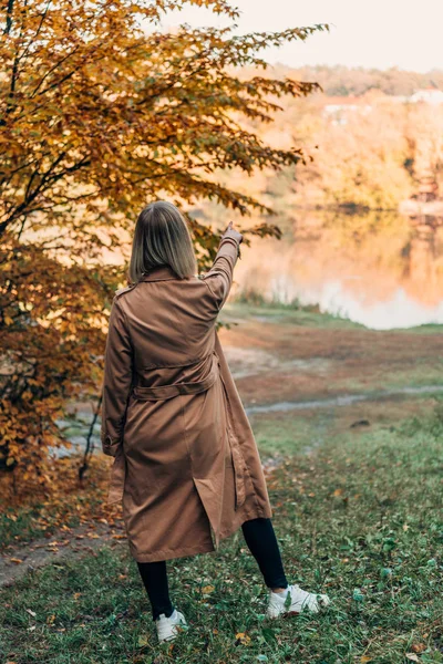 Back view of woman standing in coat and pointing at lake in autumn forest — Stock Photo