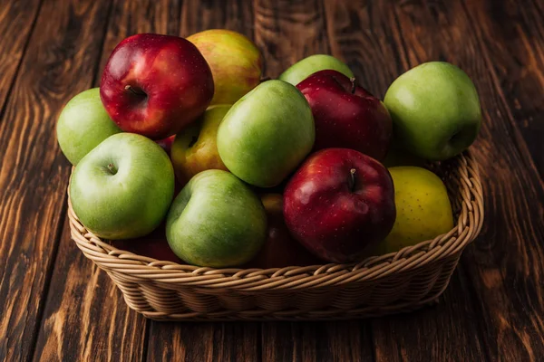 Panier en osier avec de délicieuses pommes rouges, vertes et jaunes sur table en bois — Photo de stock