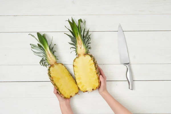 Vista recortada de manos femeninas sosteniendo mitades de piña cerca del cuchillo - foto de stock