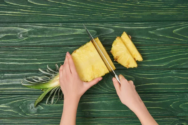 Cropped view of woman cutting yellow ripe pineapple on green wooden table — Stock Photo