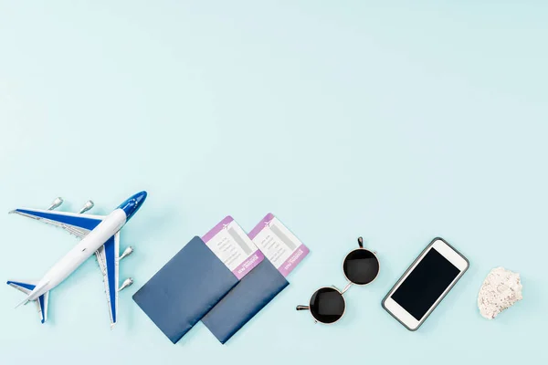 Vista superior de pasaportes, billetes de avión, teléfono inteligente avión de juguete con pantalla en blanco, conchas marinas y gafas de sol sobre fondo azul - foto de stock