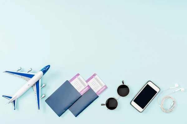 Vista superior de pasaportes, billetes de avión, avión de juguete, smartphone con pantalla en blanco, auriculares y gafas de sol sobre fondo azul - foto de stock
