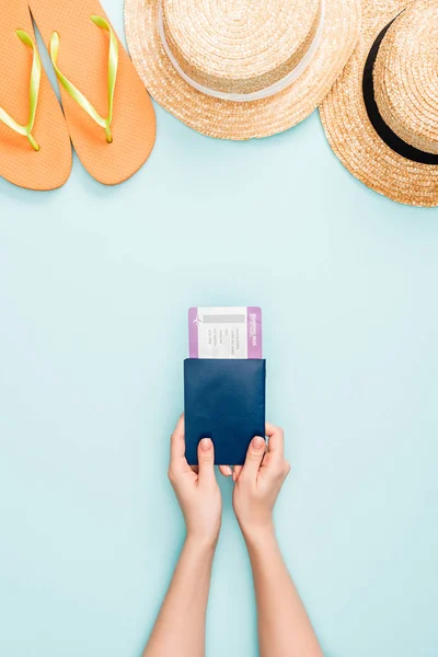 Vista ritagliata di donna in possesso di passaporto con biglietto aereo vicino cappelli di paglia e infradito su sfondo blu — Foto stock