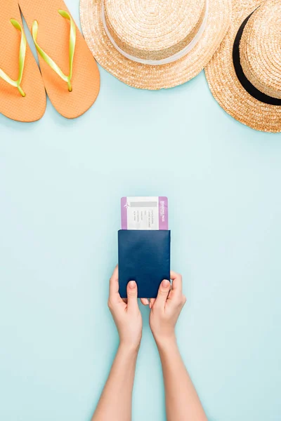 Vista recortada de la mujer con pasaporte con billete de avión cerca de sombreros de paja y chanclas sobre fondo azul - foto de stock