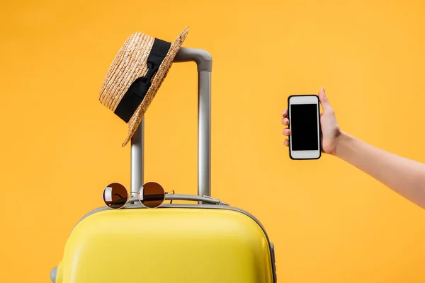 Vista recortada de la mujer sosteniendo teléfono inteligente con pantalla en blanco cerca de la bolsa de viaje, sombrero de paja y gafas de sol aisladas en amarillo - foto de stock