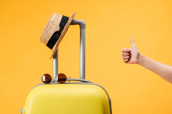 Vista recortada de la mujer haciendo gesto pulgar hacia arriba cerca de la bolsa de viaje, sombrero de paja y gafas de sol aisladas en amarillo - foto de stock