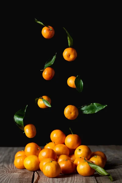 Tasty orange tangerines with green leaves falling on wooden table isolated on black — Stock Photo