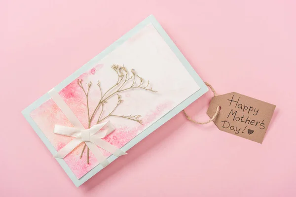 Tarjeta de felicitación decorada con planta seca, arco y etiqueta de madera con texto de saludo feliz día de las madres sobre fondo rosa - foto de stock